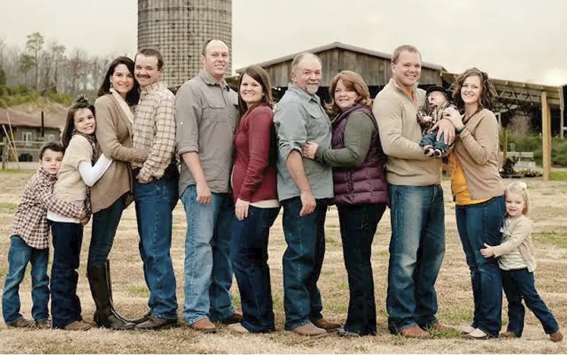Portrait of a large family posing outdoors in front of a barn and silo, with adults and children standing close together, dressed in casual winter clothing