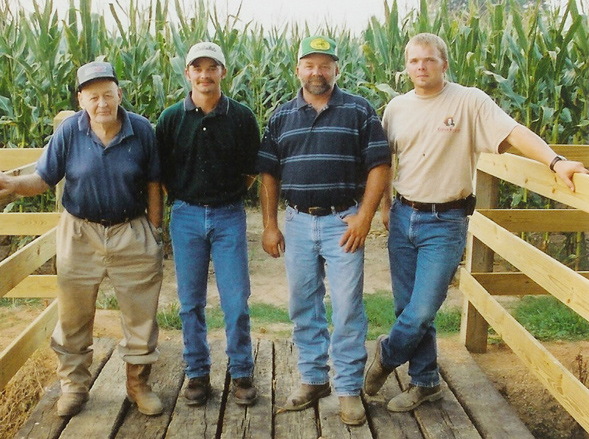 Four men standing on a wooden bridge in front of a cornfield, dressed in casual farm attire, representing different generations of a farming family.