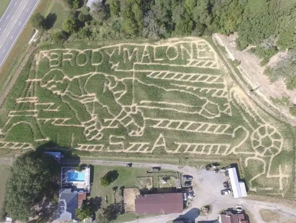 Aerial view of a corn maze featuring gymnast Brody Malone with gymnastic equipment, American flag elements, and the year '2024'.